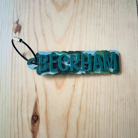 Keychain with a camouflage background and single colored name overlaid in acrylic with a wire keyring