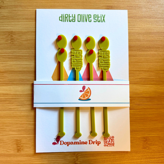 Packaging of 'Dirty Olive Stix' with colorful olive-shaped tools on a wooden surface.