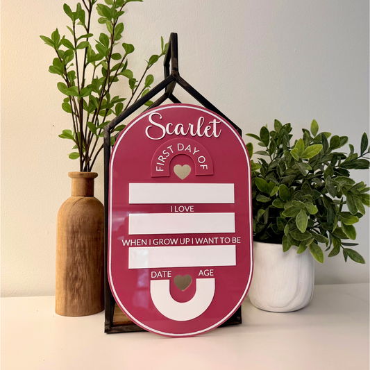 Pink milestone plaque with text and heart shapes, placed on a surface with plants in the background.