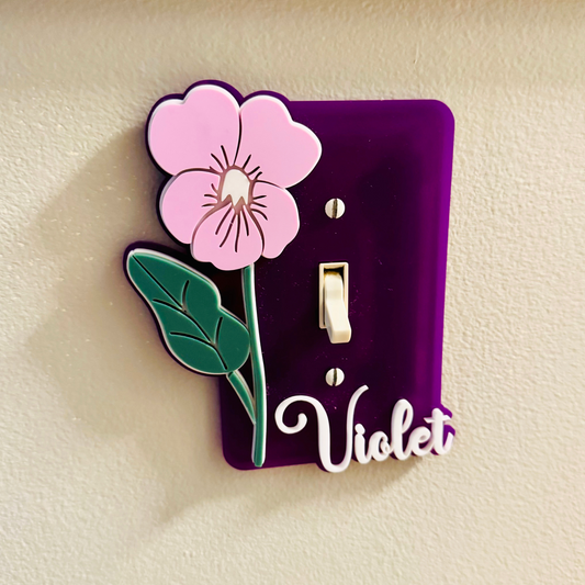 Decorative light switch cover with a pink flower and 'Violet' text on a beige background
