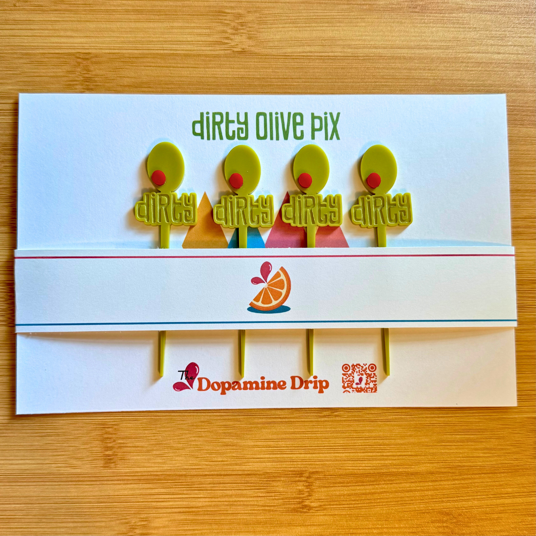 Product packaging for dirty olive cocktail picks with colorful characters and 'Dopamine Drip' branding on a wooden surface