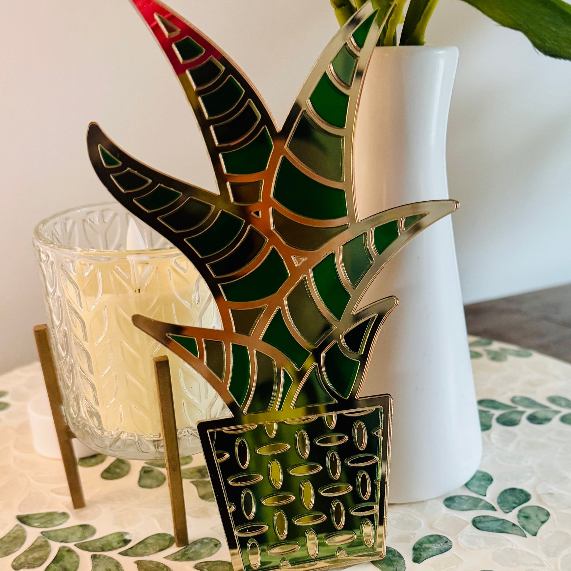 Decorative glass plant with green and red design on a table with a white vase and candle in the background.